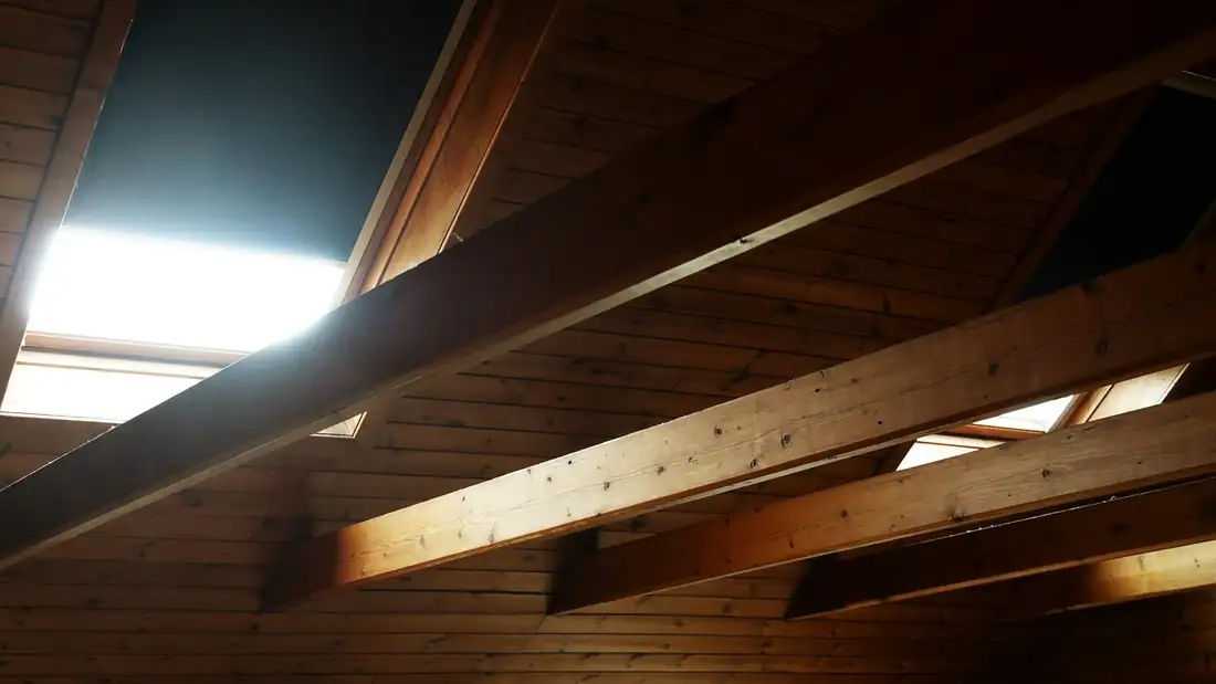 Poutres en bois sombre sous un rampant lambrissé, lumière d’une fenêtre de toit