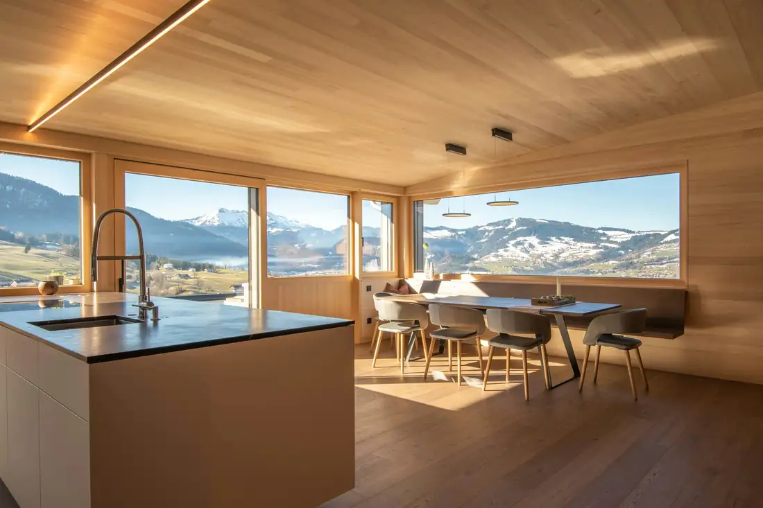 Pièce à vivre entièrement boisée, cuisine ouverte et grandes baies sur des montagnes enneigées